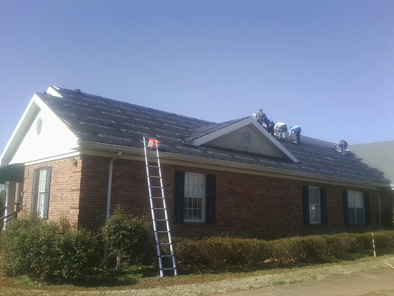 Crew tearing shingles off a brick ranch roof with a ladder set — Metal Roof Repair work by our crew in Camp Grove