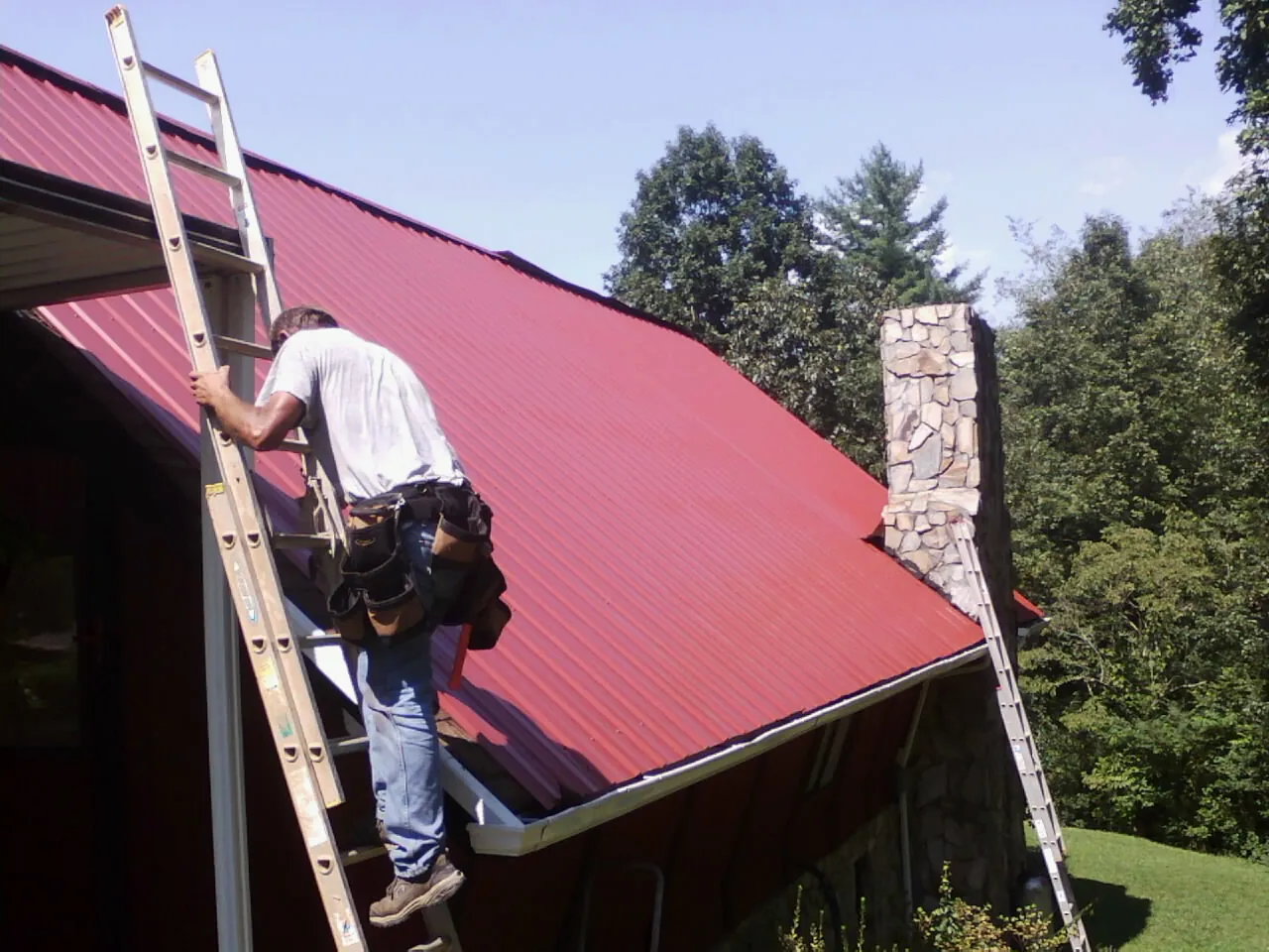 Roofer on a ladder beside a red metal roof and stone chimney — Soffit & Fascia Repair by Roofing Brothers in Camp Grove